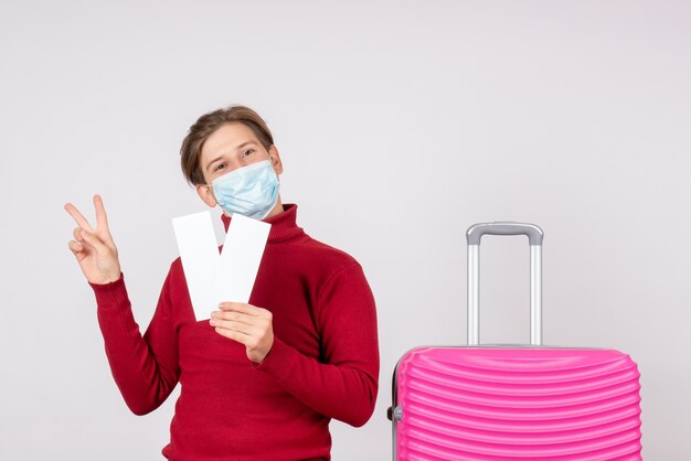 Front view of young male in mask holding plane tickets on white wall