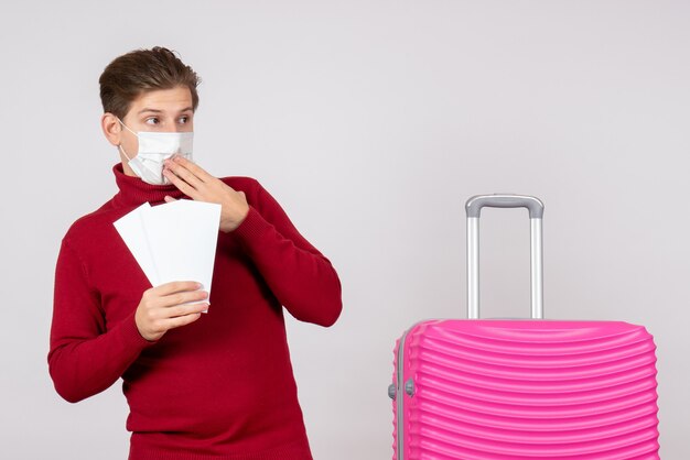 Front view of young male in mask holding plane tickets on white wall