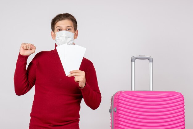 Front view of young male in mask holding plane tickets on white wall