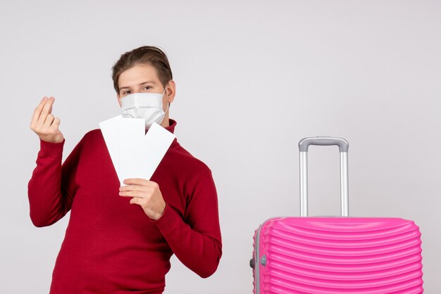 Front view of young male in mask holding plane tickets on white wall