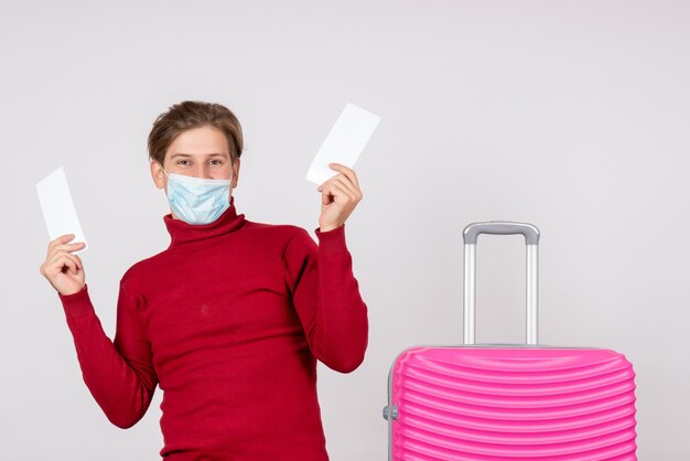 Front view of young male in mask holding plane tickets on white wall