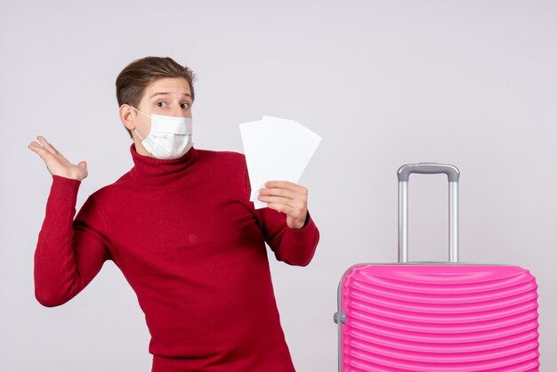 Front view of young male in mask holding tickets on white wall