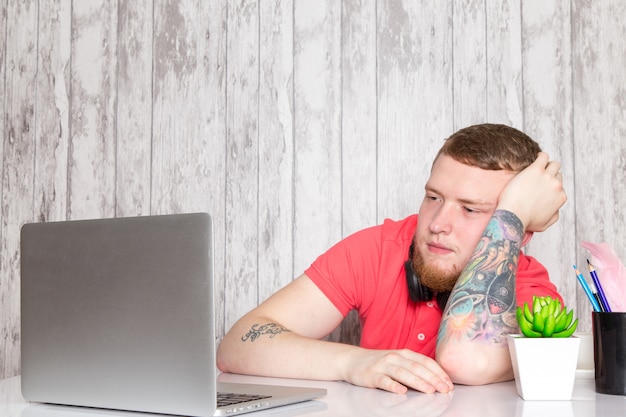 A front view young male in pink shirt in black earphones using grey laptop table with pens coffee plant grey space