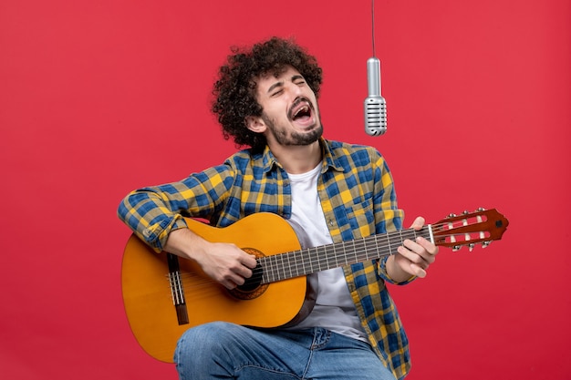 Вид спереди молодой мужчина играет на гитаре и поет на красной стене, выступление группы музыкант, концерт, живая цветная музыка