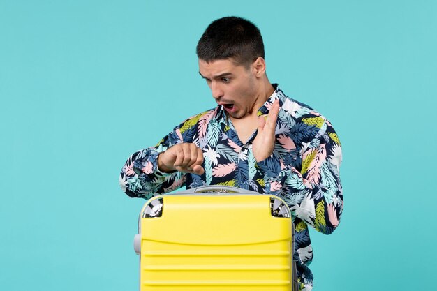 Front view of young male preparing for trip with his yellow bag and checking time on the blue wall