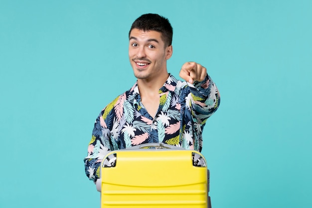 Front view of young male preparing for trip with his yellow bag pointing out on blue wall