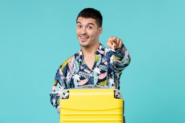 Front view of young male preparing for trip with his yellow bag pointing out on blue wall