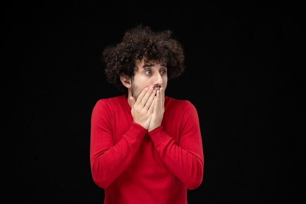 Front view of young male in red sweater scared on black wall