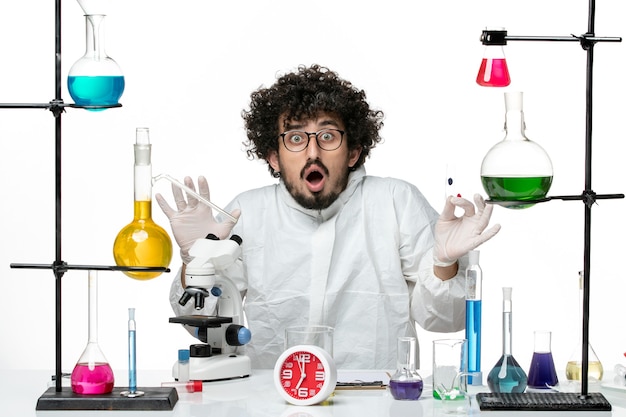 Вид спереди молодой ученый-мужчина в специальном костюме, держащий образцы и использующий микроскоп на светлой белой стене