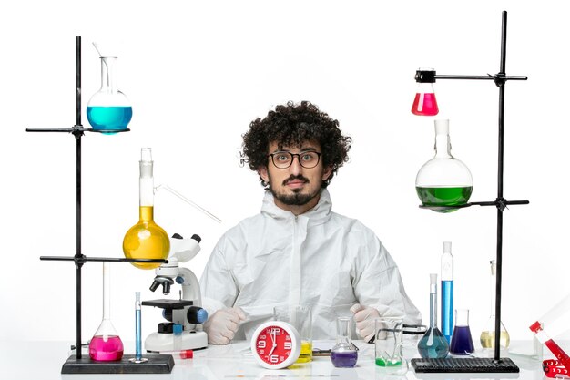 Front view young male scientist in special suit sitting with different solutions on white wall