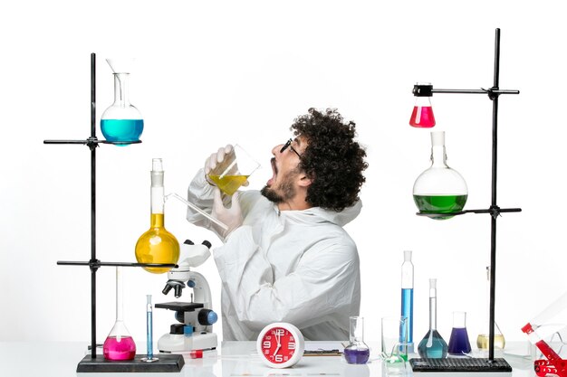 Вид спереди молодого ученого-мужчины в специальном костюме, сидящего с растворами на белой стене