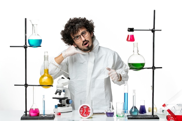 Vista frontale giovane scienziato maschio in abito speciale in piedi intorno al tavolo con soluzioni su un muro bianco chiaro science lab covid- chimica pandemica