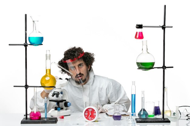 Front view young male scientist in special suit with protective helmet using microscope on a white wall science lab covid chemistry male