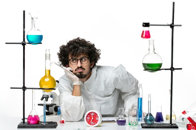 Front view young male scientist in white special suit sitting around table with solutions on white wall science covid pandemic chemistry lab