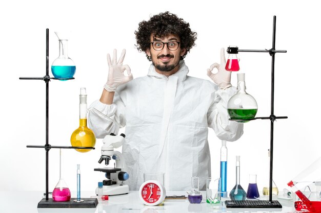 Front view young male scientist in white special suit standing around table with solutions smiling