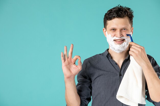 正面図青い背景にかみそりで彼の泡の顔を剃っている若い男性