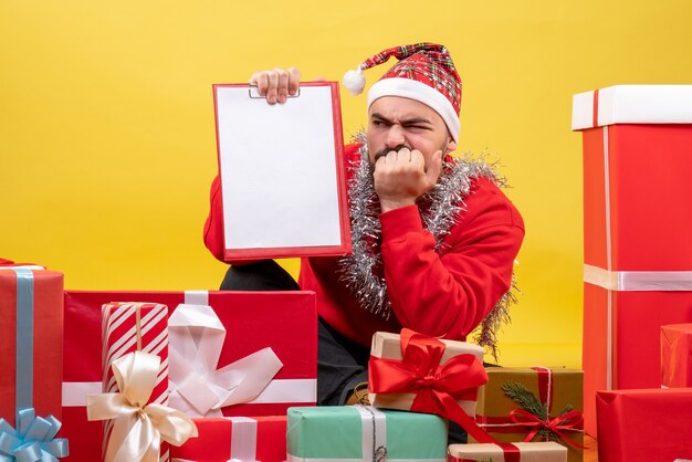 Вид спереди молодого мужчины, сидящего вокруг рождественских подарков с запиской на желтом фоне