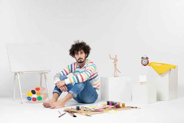 Front view of young male sitting around paints and drawings bored on white wall