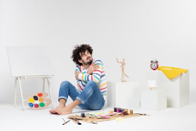 Вид спереди молодого мужчины, сидящего вокруг красок и рисунков, дрожащего на белой стене