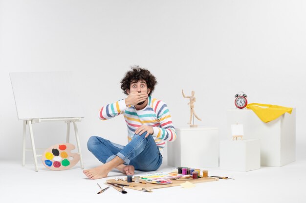 Front view of young male sitting around paints and drawings surprised on white wall