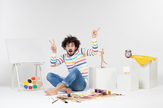 Front view of young male sitting around paints and drawings on white wall