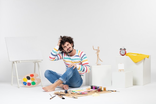 Front view of young male sitting around paints and drawings on white wall