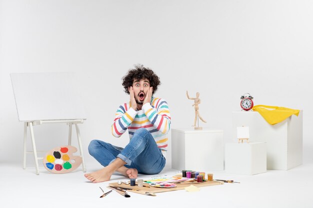Free photo front view of young male sitting around paints and drawings on white wall