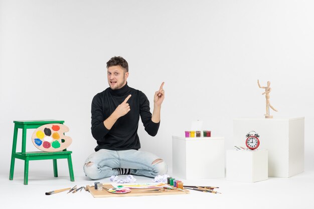 Вид спереди молодой мужчина сидит с красками и кисточками для рисования на белой стене искусство рисовать краской живопись цвет художник