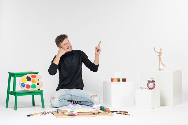 白い壁に描くための絵の具やタッセルの周りに座っている正面図若い男性カラー絵の具アート絵画アーティストが描く