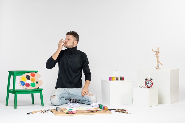 Vista frontale giovane maschio seduto intorno a vernici e nappe per disegnare sul muro bianco disegnare immagine a colori pittura artista pittura arte
