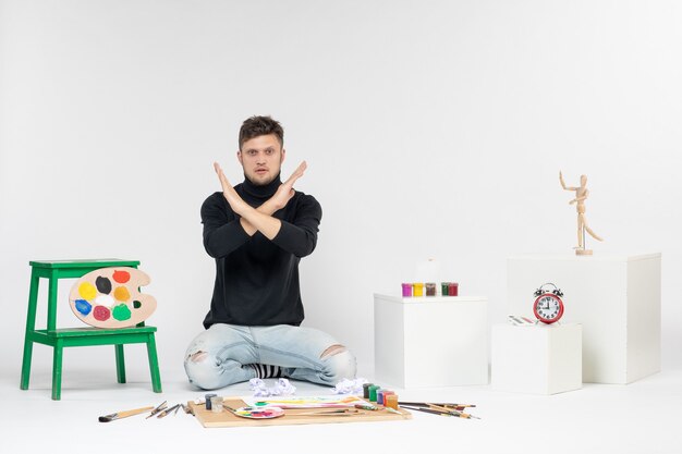 白い壁に描くための絵の具やタッセルの周りに座っている正面図の若い男性は、カラー絵の絵を描くアーティストのペイントアートを描く