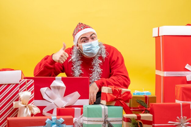 正面図黄色の背景色のパンデミックウイルスのクリスマスに無菌マスクでプレゼントの周りに座っている若い男性