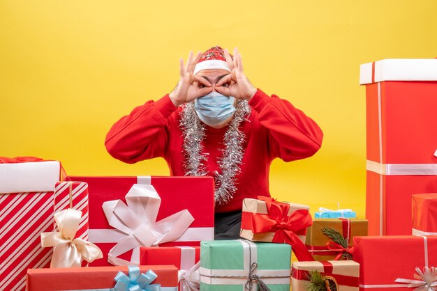 Front view young male sitting around presents in mask on the yellow background