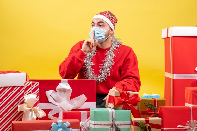 Front view young male sitting around presents in mask on the yellow background