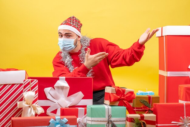 Front view young male sitting around presents in mask on the yellow background