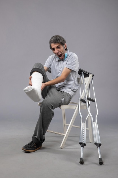 Front view young male sitting with bandaged broken foot on grey desk leg accident twist male foot pain