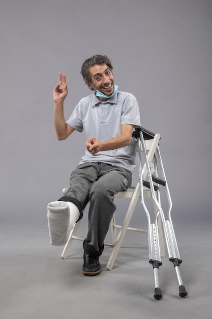 Вид спереди молодого мужчины, сидящего с перевязанной сломанной ногой на серой стене, несчастный случай с болью в мужской ноге