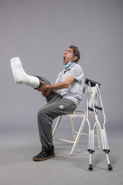 灰色の壁の痛みの足の事故のねじれ男性の足に包帯の壊れた足で座っている正面図若い男性