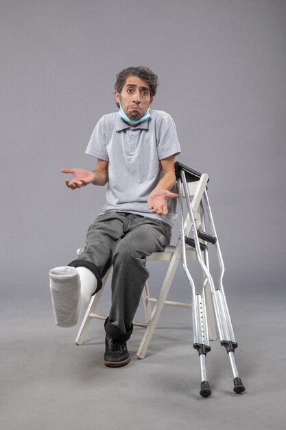 Front view young male sitting with bandaged broken foot on the grey wall pain twist leg accident male foot