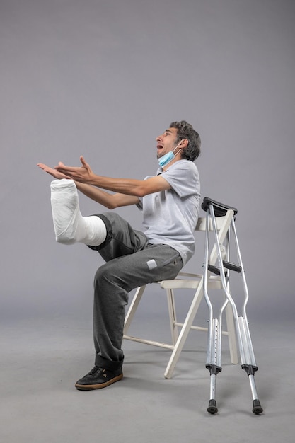 Vista frontale giovane maschio seduto con piede rotto bendato che urla dal dolore sul muro grigio dolore alla gamba incidente piede maschio torsione