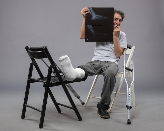 Бесплатное фото Вид спереди молодого мужчины, сидящего со сломанной ногой и держащего рентгеновский снимок на серой стене, скручивающей боль в ноге, сломанной ногой несчастного мужчины