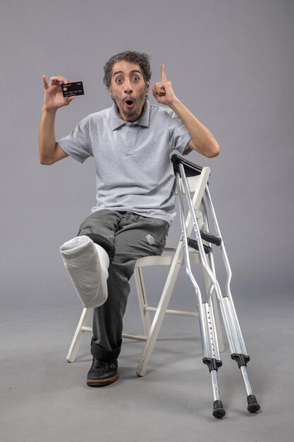 Front view young male sitting with broken foot and bandage holding bank card on grey wall male accident twist foot pain