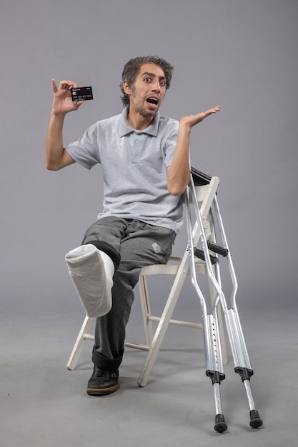 Вид спереди молодого мужчины, сидящего со сломанной ногой и повязкой, держащего банковскую карту на серой стене, поворот мужской аварии, боль в ноге, нога