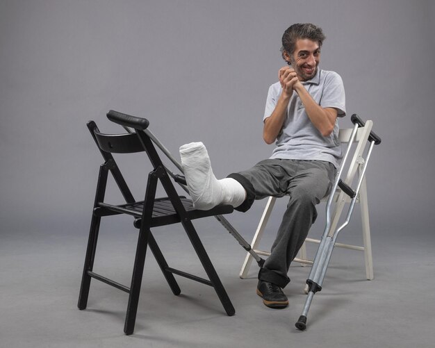 正面図灰色の壁の事故で幸せを感じて足の骨折と松葉杖で座っている若い男性ねじれ脚の痛み足の骨折