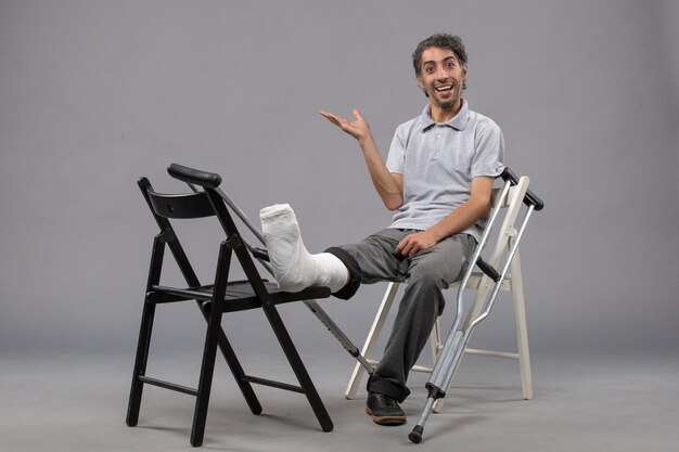 Front view young male sitting with broken foot and crutches on grey desk accident leg broken twist foot pain
