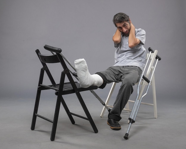 灰色の机の事故で足の骨折と松葉杖で座っている正面図若い男性が足の骨折した足の痛みをひねる