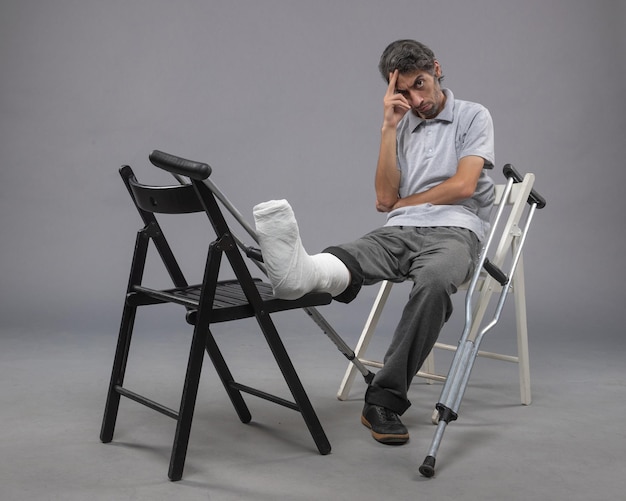 Front view young male sitting with broken foot and crutches on grey floor broken accident foot legs pain twist