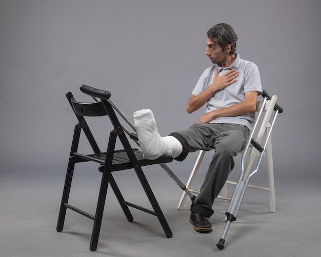 Free photo front view young male sitting with broken foot and crutches on grey floor pain accident twist foot leg broken