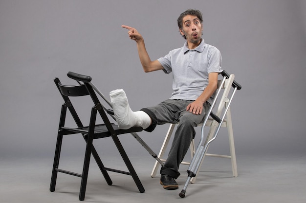 Front view young male sitting with broken foot and crutches on grey wall accident broken twist foot pain leg