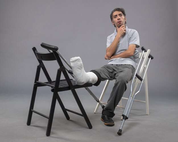 Front view young male sitting with broken foot and crutches on grey wall broken accident foot leg pain twist